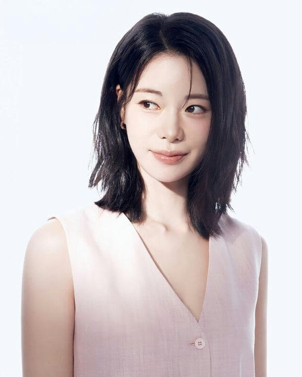 This image is a portrait photograph of a young East Asian woman, likely a Korean celebrity or model, given the style. Here is a detailed description of what is in the image: * **Subject:** A woman with fair skin and dark, shoulder-length hair styled in a slightly layered or choppy cut. Her hair is parted slightly off-center. * **Facial Expression & Pose:** She is looking slightly off-camera to the viewer's left with a soft, gentle expression and a slight, closed-mouth smile. Her makeup appears natural and dewy. * **Attire:** She is wearing a sleeveless top or vest in a very light pink or blush color. The garment features a deep V-neckline and appears to have a single visible button near the center bottom of the frame. The fabric has a subtle texture. * **Background:** The background is uniformly bright white or very light gray, suggesting a studio setting with bright, high-key lighting that emphasizes the subject. * **Lighting:** The lighting is bright and flattering, creating subtle shadows that define her features and highlights on her skin and hair.