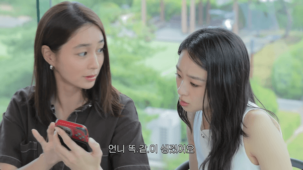 people-Two young East Asian women-are indoors-looking at a red object (possibly a phone or case) held by the woman on the left-Korean text overlay reads 