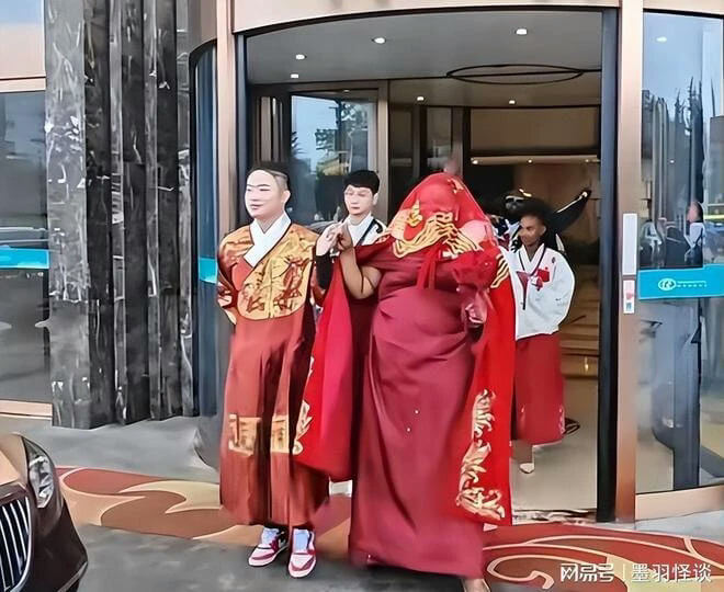 people-Two individuals in traditional/historical-style Chinese garments-A-man in maroon/red robe with gold embroidery and sneakers-A-woman covered mostly by a flowing red veil/shawl with gold trim-A-people standing in a doorway of a modern building, possibly exiting-B-person in white and red attire behind the main figures-C-car visible on the left edge