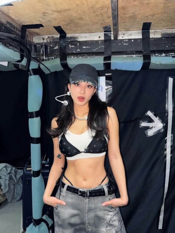 people-One young woman-is wearing-a black baseball cap-a black and white bikini top layered over a white sports bra-grey, low-rise jeans with a visible black thong strap and belt-she has long dark hair and is posing indoors against a dark background.