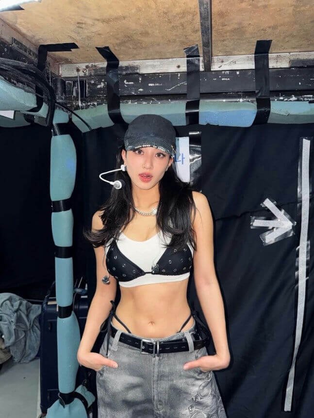 people-One young woman-is wearing-a black baseball cap-a black and white bikini top layered over a white sports bra-grey, low-rise jeans with a visible black thong strap and belt-she has long dark hair and is posing indoors against a dark background.