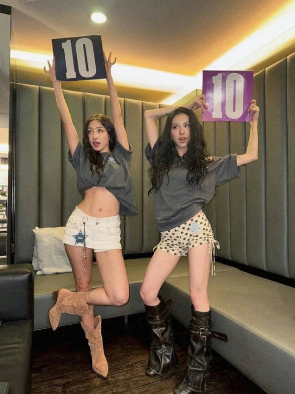 people-Two women-A-holding up scorecards-A-showing the number 10-B-wearing casual, cropped tops and short shorts-C-posing indoors with dark, textured seating in the background