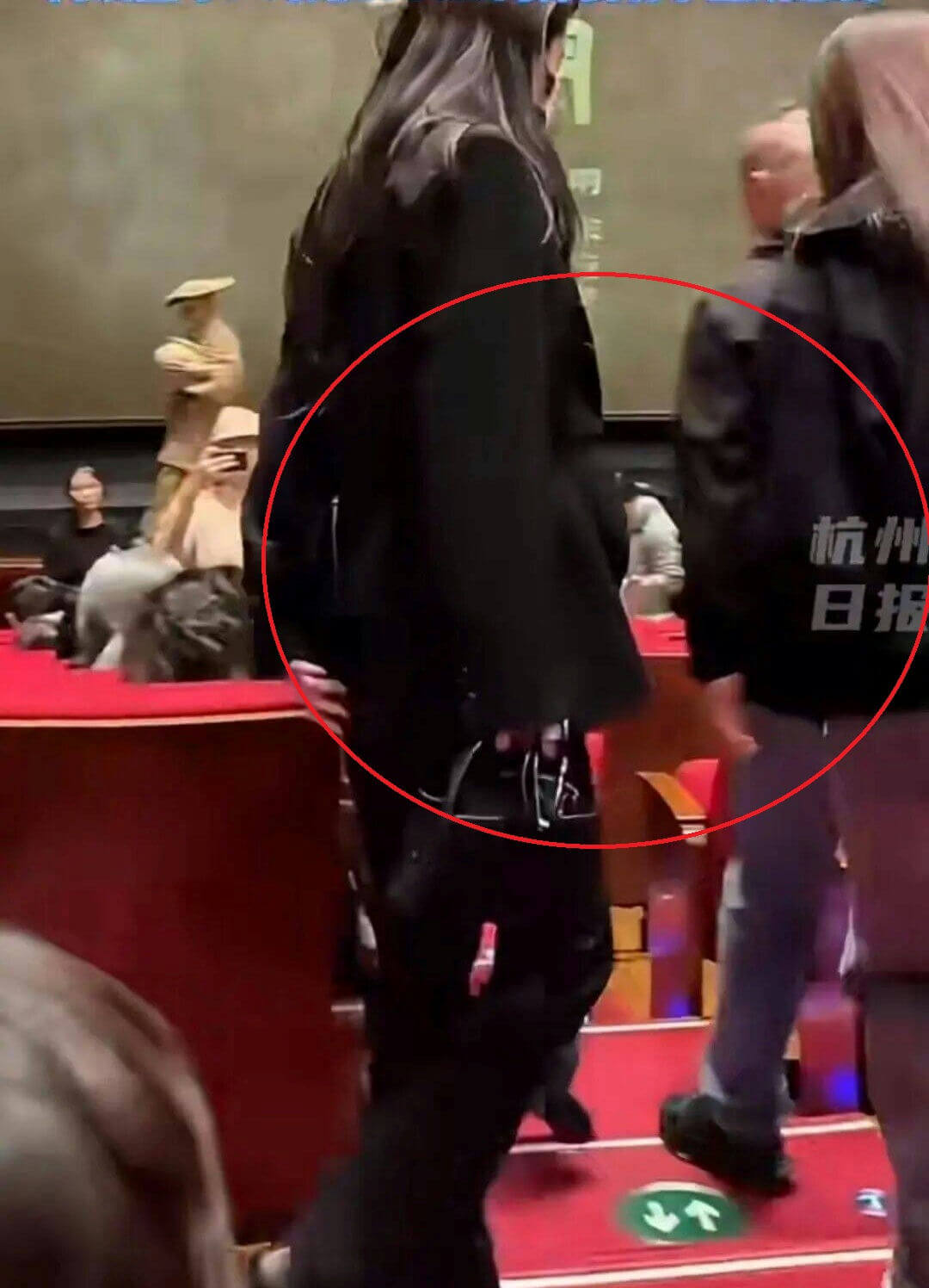 people-A-woman in black long hair and dark clothing walking-A-audience seating area with red carpeted steps-A-person in black jacket and light pants walking to the right-A-text overlay partially visible 