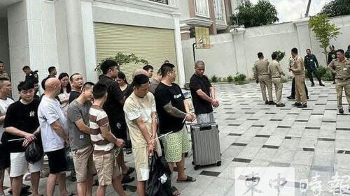 people-many men and a few women-A-being detained or arrested-A-uniformed officers present-A-tied wrists/cuffed-A-outdoor paved area-B-luggage present-B-residential or office building in background-C-Asian descent individuals visible-C