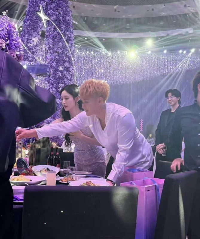 people-man with blonde hair-reaching over table-woman in sparkly dress-standing next to man-man in black suit-standing in background-event setting