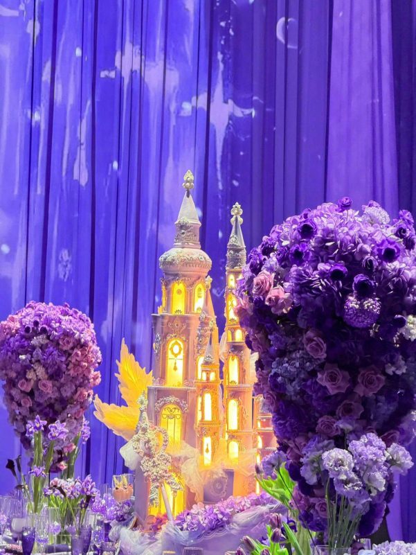 A lavishly decorated event setting featuring a multi-tiered, illuminated castle cake centerpiece, surrounded by massive, dense bouquets of purple and lavender flowers, set against a backdrop of draped, deep purple fabric with faint projections of stars or celestial bodies. people-N/A-castle cake-A-purple flowers-A-event decor-A-lavender tones-B-illuminated-B-draped fabric-C-extravagant-C