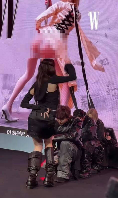 people-woman in black outfit-A-standing on stage-A-people in black leather outfits-A-seated on stage-A-large screen display in background-A-pixelated section on screen-A-woman with long dark hair-A-tall black boots-A-man with brown hair crouching-A-Korean text visible-A-W logo on screen