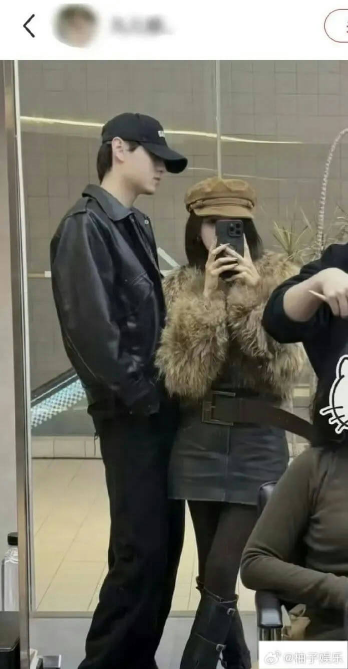 people-two people posing for a mirror selfie-man wearing a black leather jacket and black cap-woman wearing a brown fur jacket, baker boy cap, and black skirt-third person partially visible on the right
