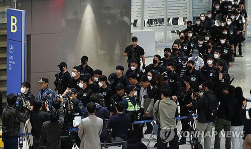 people-Many individuals, mostly men-A-Mass arrival or escort-A-Police or security personnel visible-A-Masked faces and dark clothing dominate-B-Media presence with cameras and microphones-B-Airport or large public terminal setting-C-Formal event or high-profile transport of persons-C
