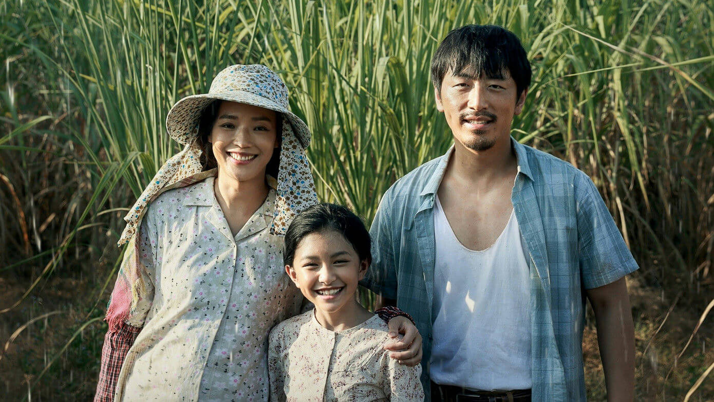 people-Three people (a woman, a man, and a young girl)-A-Smiling-A-Standing in front of tall green grass (likely sugarcane)-B-Wearing simple, light-colored, patterned clothing-C-Outdoor, sunny lighting