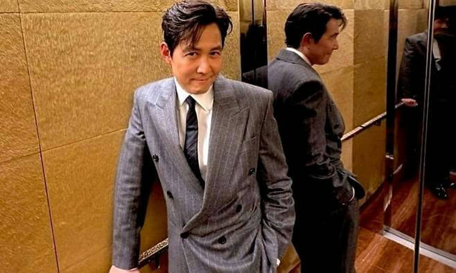 people-Korean actor Lee Jung-jae-A-wearing a grey pinstripe double-breasted suit-A-standing in what appears to be an elevator or hallway with golden-brown textured walls-A-reflected in a nearby mirror-B-subtly smiling at the camera-C