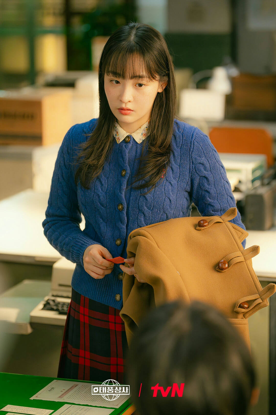people-young woman-blue cable knit cardigan-floral collar shirt-red and black plaid skirt-holding small red paper-brown duffle coat with toggles-freckles-long dark hair-tvn logo
