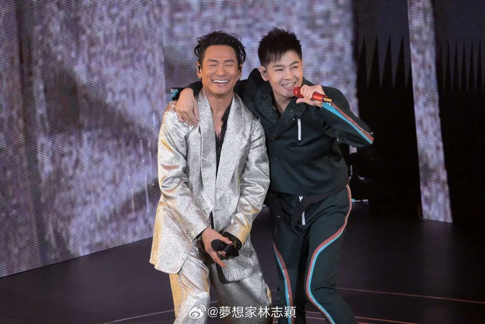 people-Two men-A-performing on stage-A-smiling-A-wearing sparkly silver suit-B-holding a microphone-B-wearing dark tracksuit with colored stripes-C-putting arm around other man