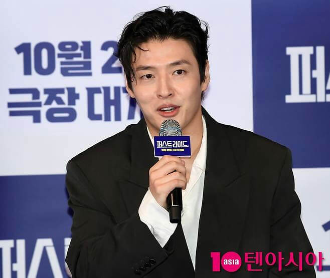 people-A-man-A-Korean actor-A-holding microphone-A-formal attire-B-press conference-B-blue background-C-text visible