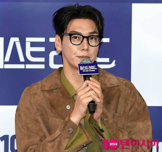 people-man-glasses-brown jacket-holding microphone-speaking-blue background