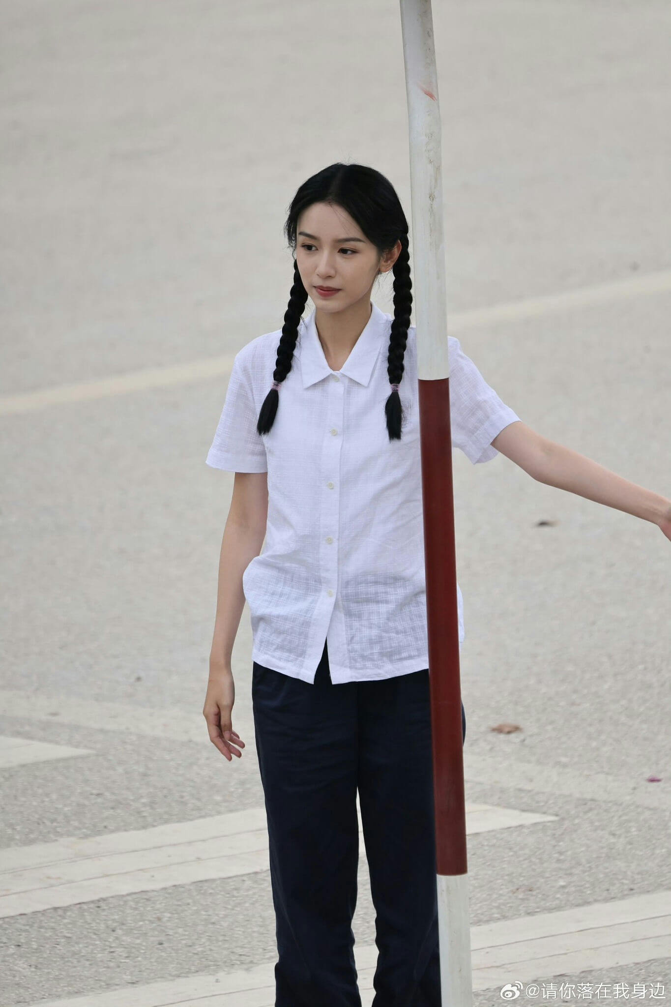 people-young woman-pigtails-white short-sleeve collared shirt-dark trousers-holding onto pole-outdoor pavement setting