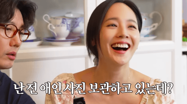 people-Woman laughing heartily while looking to the side-Man with glasses looking intently at something off-camera-background-shelves with teacups-text overlay-Korean text saying 