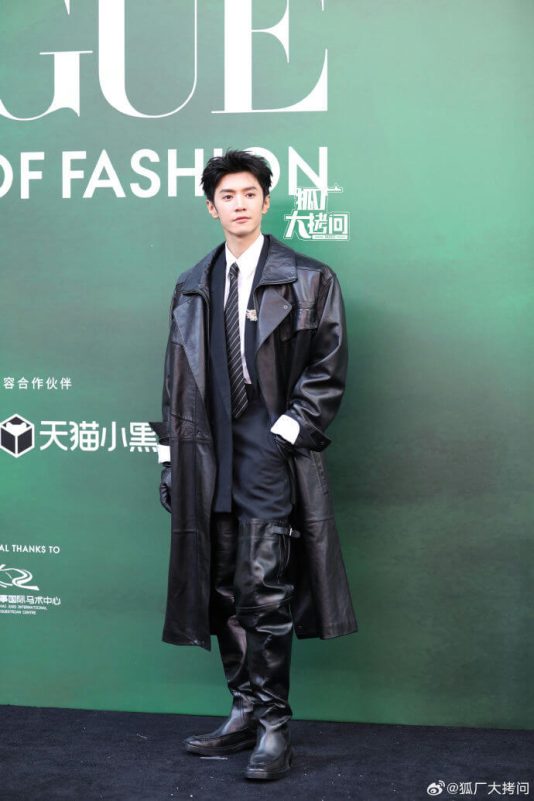 people-young man-attire-long black leather trench coat-footwear-over-the-knee black leather boots-setting-green backdrop with white text and logos
