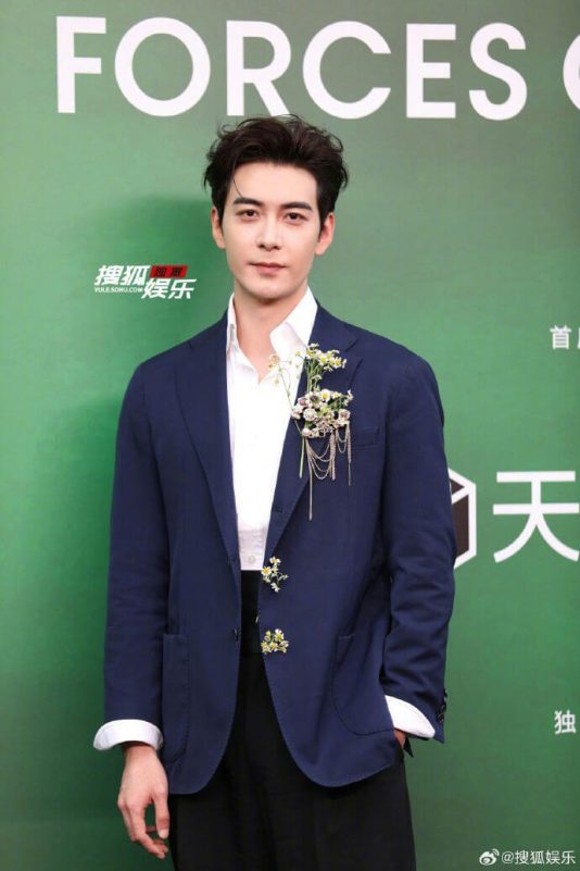 people-man-Asian-A-standing-A-formal attire-A-dark blue blazer-A-white shirt-A-black pants-A-floral lapel pin-A-green backdrop-A-text in background