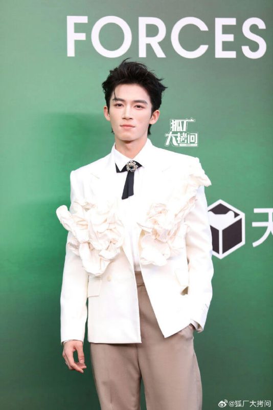people-male celebrity-A-white ruffled blazer-B-black tie with brooch-C-tan trousers