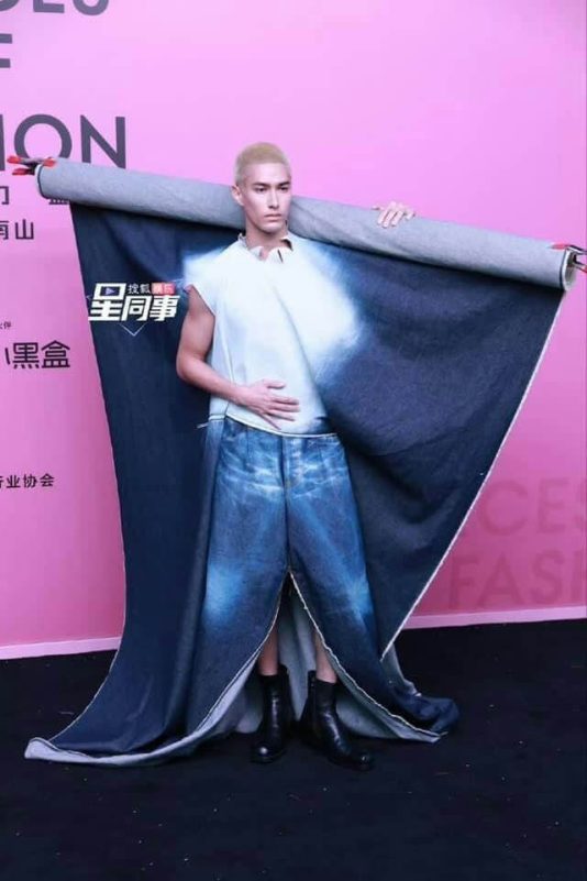 person-A-man with bleached blonde hair-A-wearing a white sleeveless top-A-wearing very wide, distressed denim trousers-A-holding a massive roll of dark blue/indigo denim fabric over his shoulders