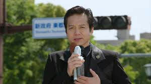 people-A-man speaking into microphone-A-outdoors setting-B-black suit jacket-C-Japanese text sign in background