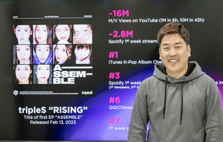 people-A-man-A-smiling-B-wearing-grey-hoodie-C-standing-in-front-of-a-screen-displaying-K-pop-album-achievements