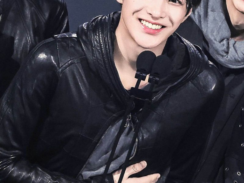 people-young man-A-smiling brightly-A-wearing a black leather hooded jacket-A-holding his hand over his stomach/chest area-A-surrounded by others on stage (partially visible)-A-holding lapel microphones