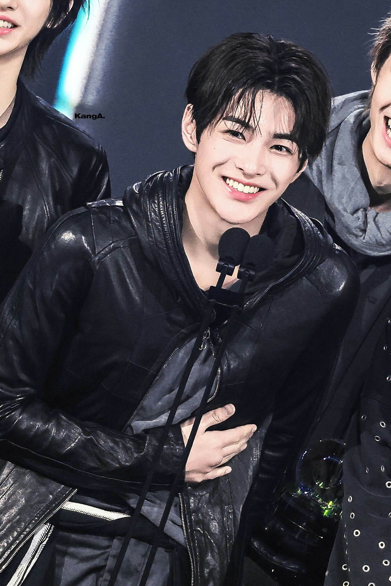 people-young man-A-smiling brightly-A-wearing a black leather hooded jacket-A-holding his hand over his stomach/chest area-A-surrounded by others on stage (partially visible)-A-holding lapel microphones