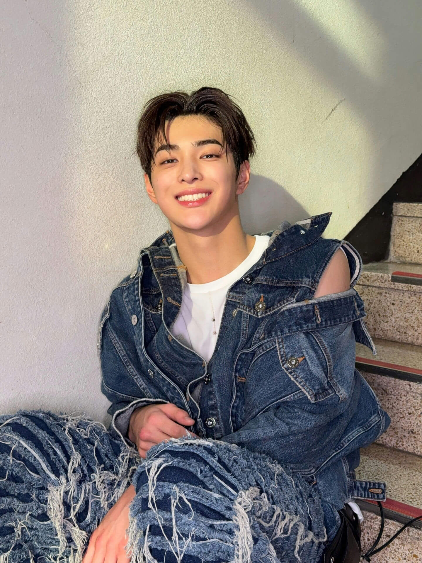 people-young man-smiling-A-wearing distressed denim jacket and ripped jeans-B-sitting indoors near a textured wall and stairs-C-dark hair
