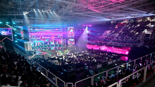 A wide shot captures a large indoor arena set up for an event, likely an awards show or presentation, featuring a brightly lit stage with large LED screens displaying graphics that include the text