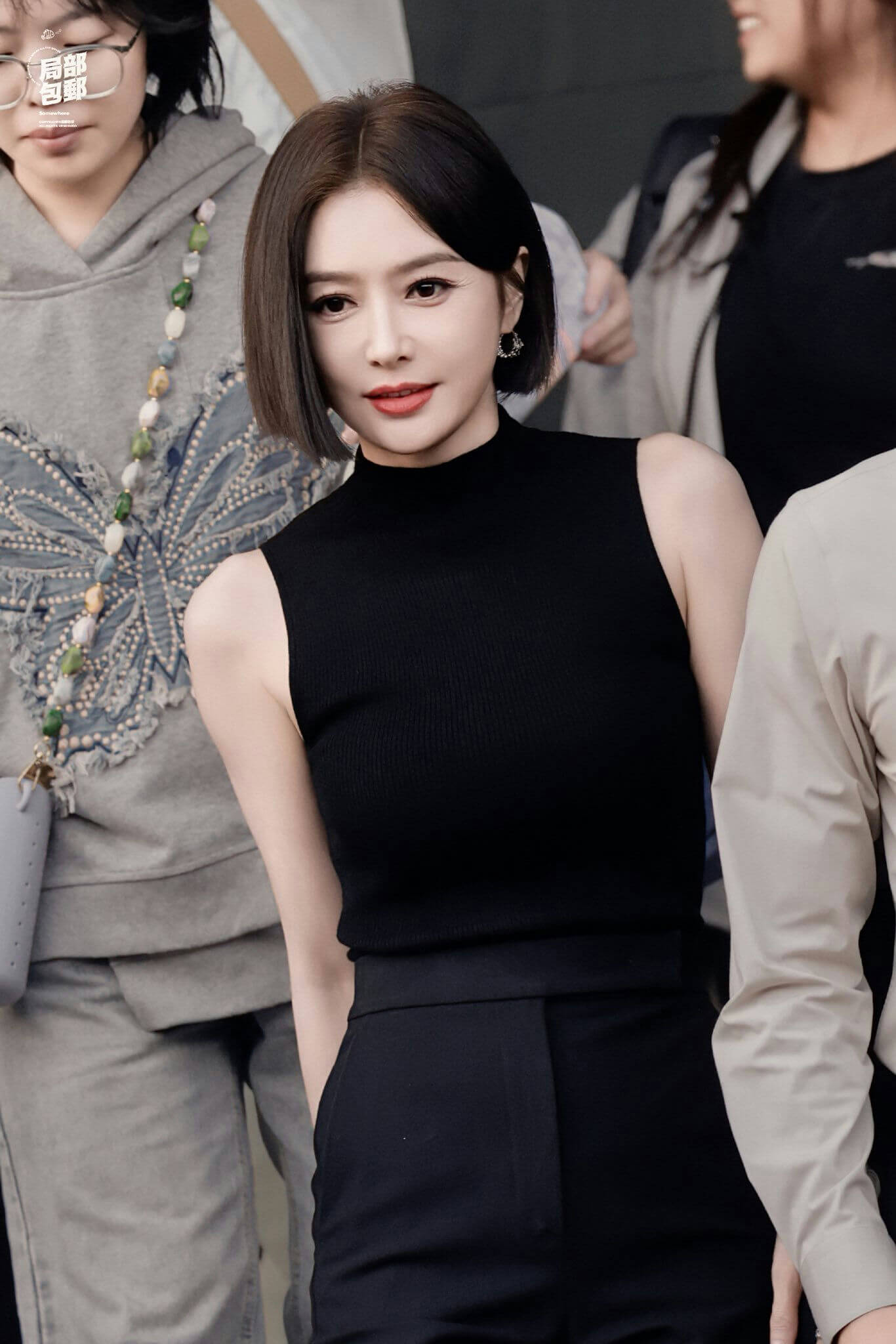 people-woman in black top-A-short black hair-A-looking at camera-A-outdoor setting-A-casual crowd background-A-jewelry earrings-A-formal pants-A-red lipstick-A-fashionable-A-celebrity-A