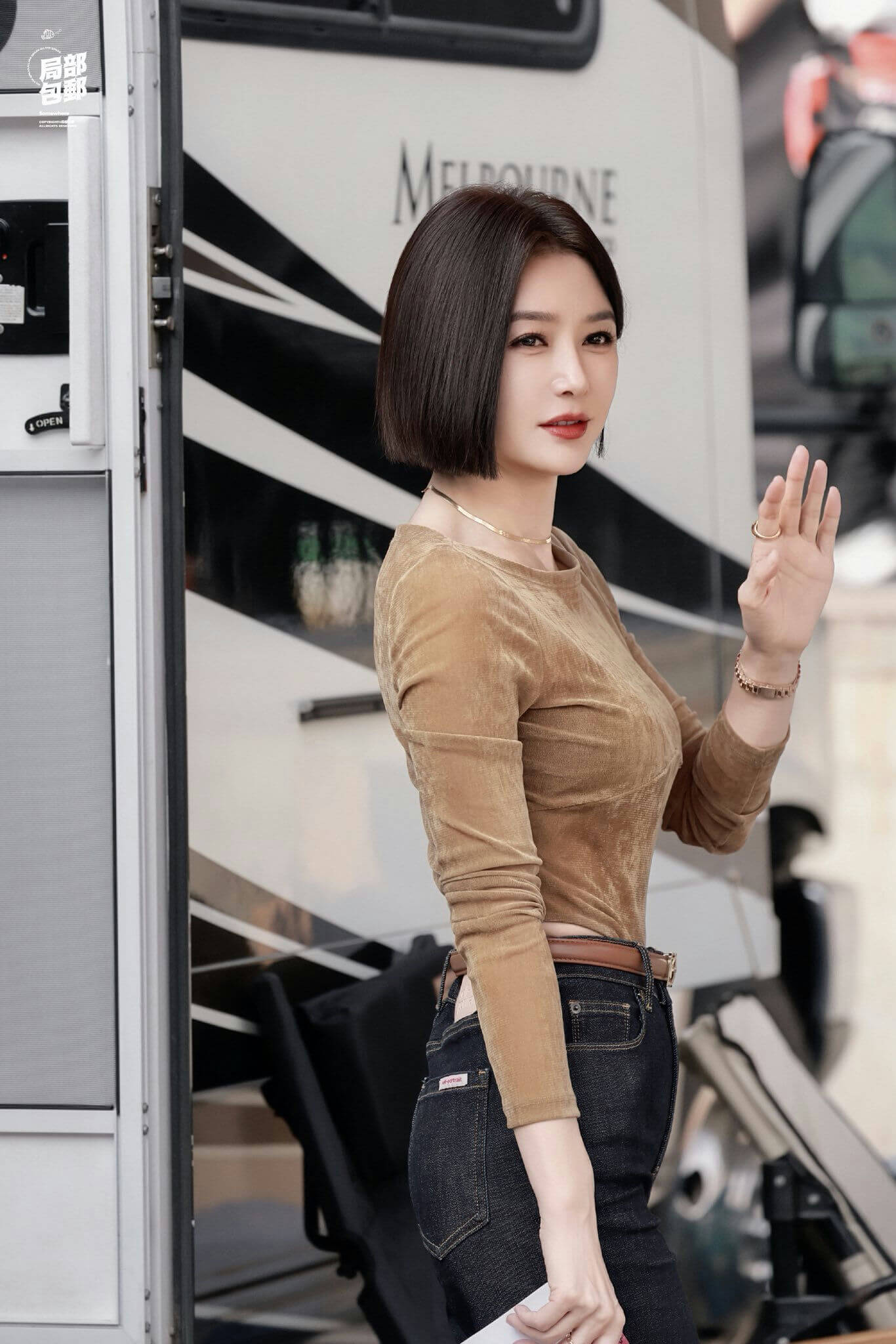 people-A-woman-A-short brown bob haircut-A-waving hand-A-brown velvet long-sleeve top-A-dark denim jeans-A-standing outdoors in front of an RV or trailer