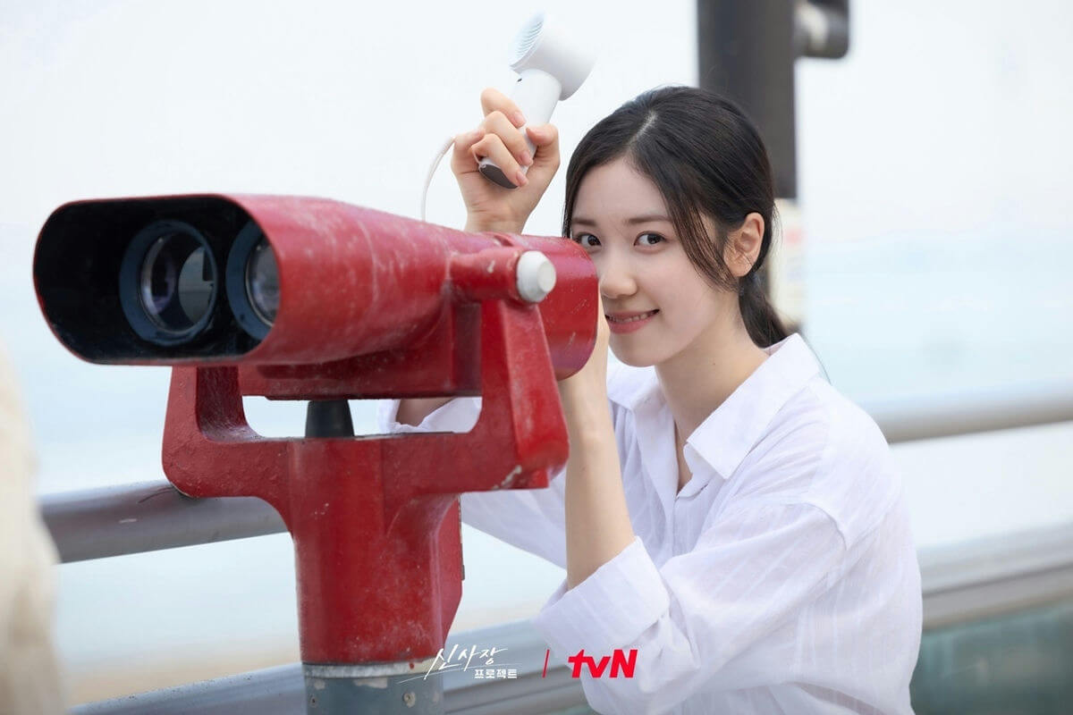 people-A-young woman-A-smiling-A-looking through binoculars-A-holding a small white fan-A-wearing a white shirt-B-red coin-operated binoculars-B-outdoor setting-C-Korean drama promotional image (tvN logo visible)