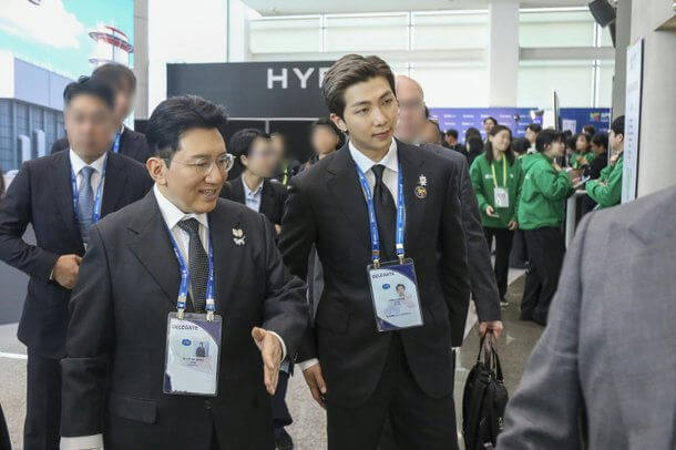people-RM (BTS)-A-formal suit-A-delegate badge-A-event venue-B-older man-B-smiling-C-staff in green shirts