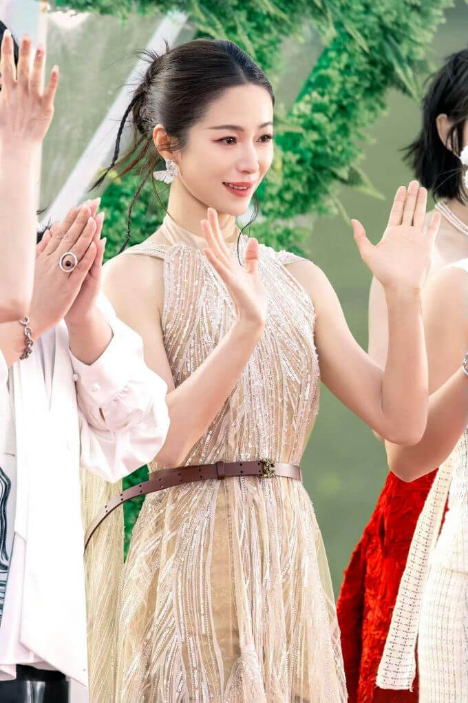 people-woman in beige sequined dress-waving-smiling-butterfly earrings-brown belt-green foliage background
