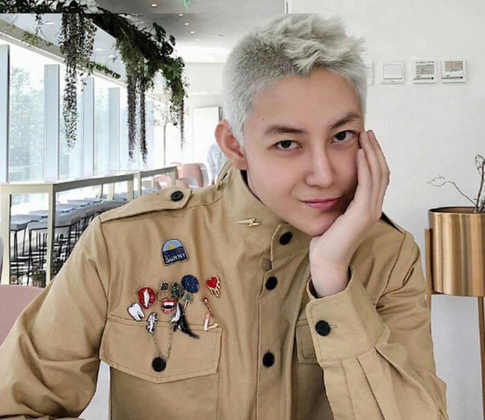 people-young man-A-platinum blonde hair-A-khaki jacket with pins-A-resting chin on hand-A-indoor setting