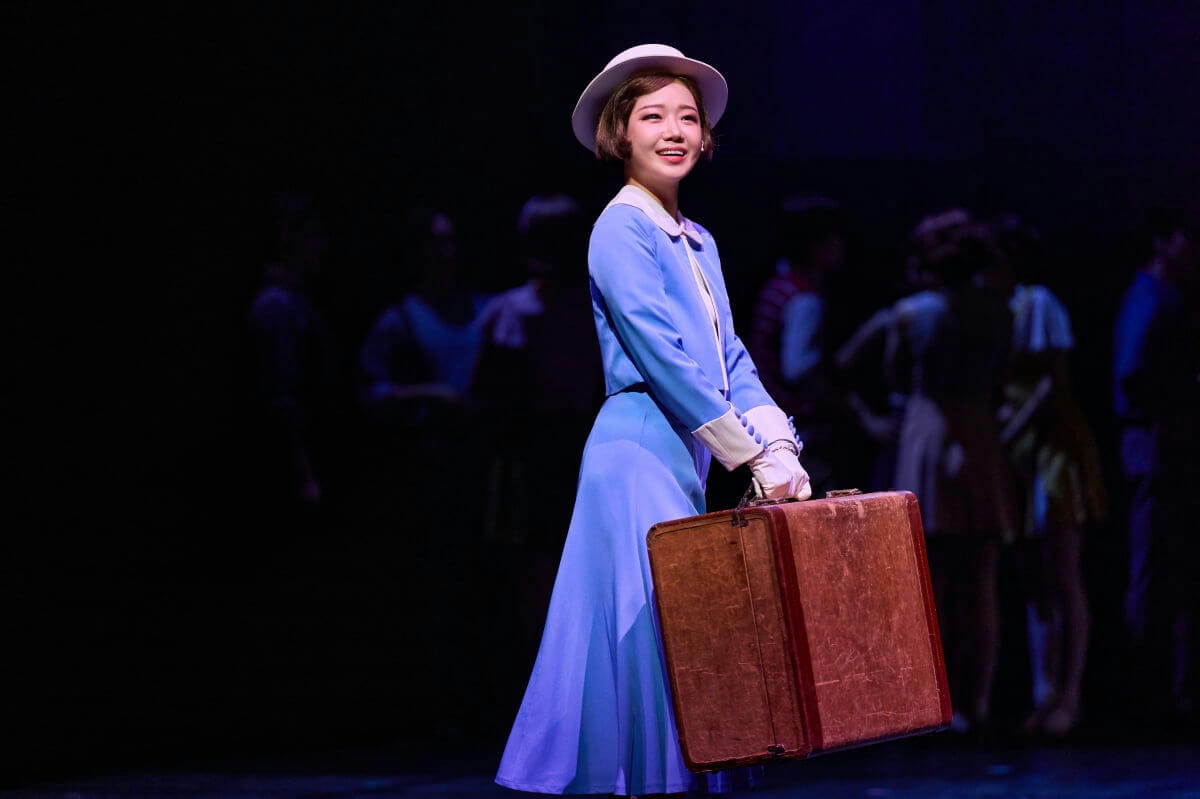 people-A-woman-A-performer-A-blue-dress-B-white-hat-B-carrying-suitcase-C-stage-C-smiling