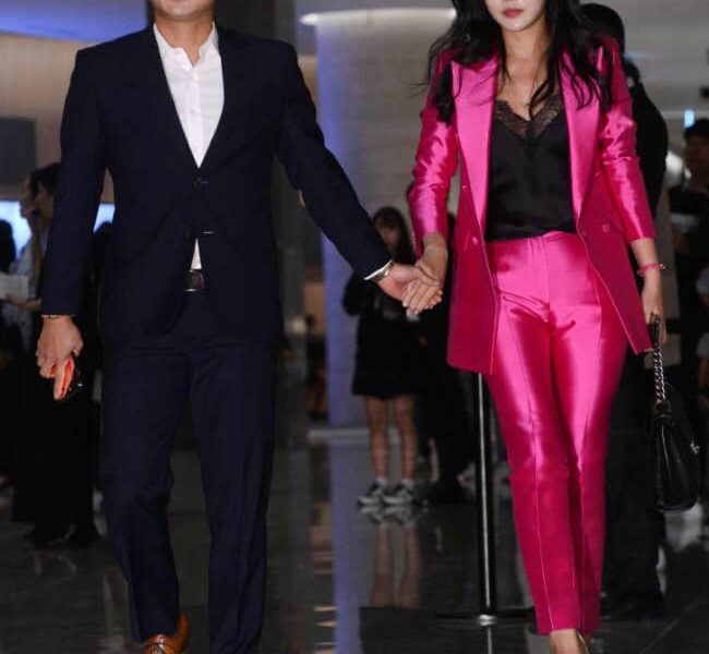 people-A-man in dark suit-A-woman in pink suit-A-couple holding hands-B-walking indoors-B-shiny floor-C-event entrance