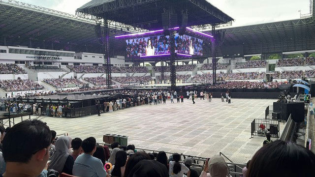 people-many people in stadium seats and on the floor-event-A-concert or performance-B-large video screen showing a woman-C-stadium seating structure