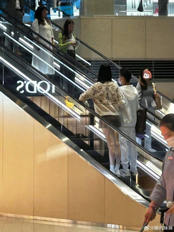 people-four women and one man on an escalator-two women standing near the top-one woman in a white dress-one woman in a green cardigan and dark top-one woman wearing a patterned sweater and sheer bottom-one man in a white hoodie and face mask-one person in a uniform cleaning the floor