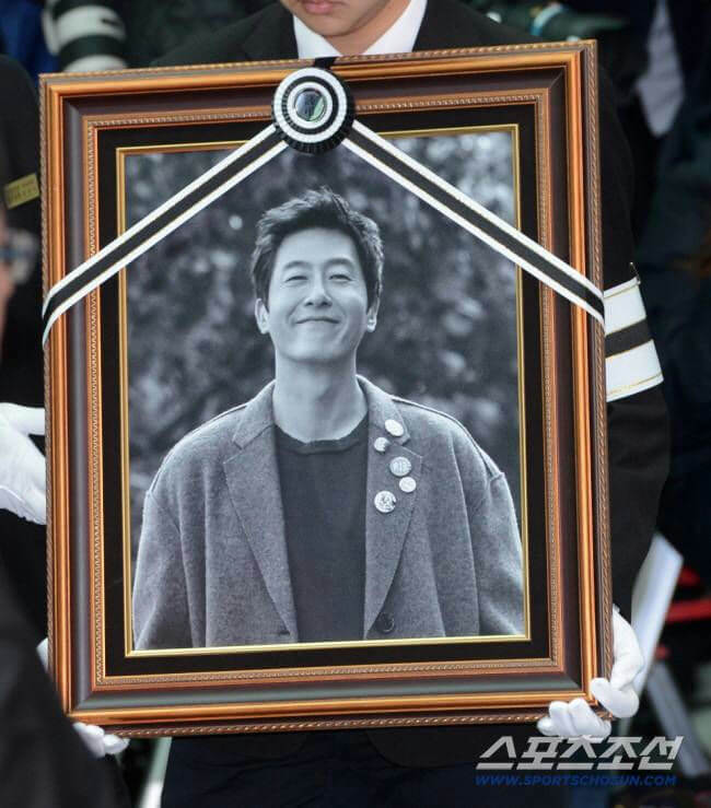 people-A-man in framed black and white photo smiling-A-funeral banner with black and white ribbon-B-person in black suit holding photo with white gloves-C-memorial portrait