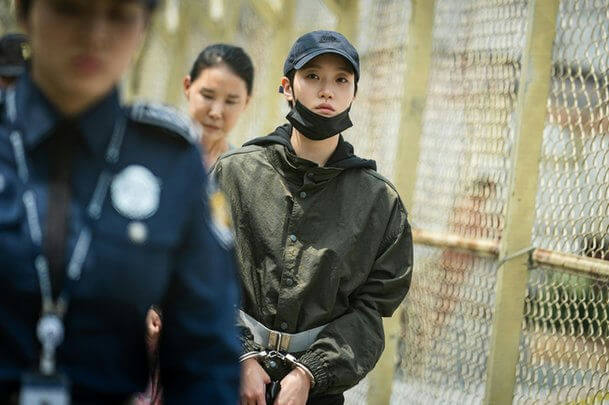people-young person in handcuffs and face mask-A-police officer in uniform-A-woman looking on-A-chain link fence background
