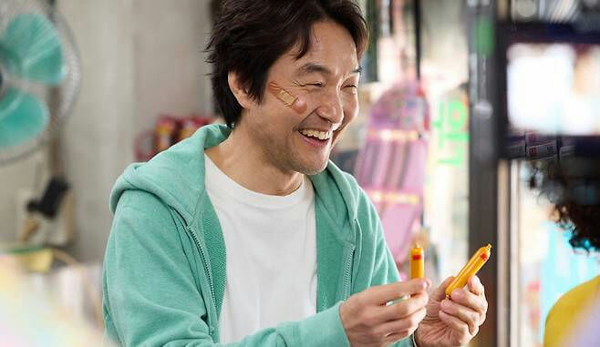 people-One man laughing joyfully-A band-aid on his cheek-B holding two yellow candy-like objects-C wearing a teal hooded sweatshirt and white t-shirt