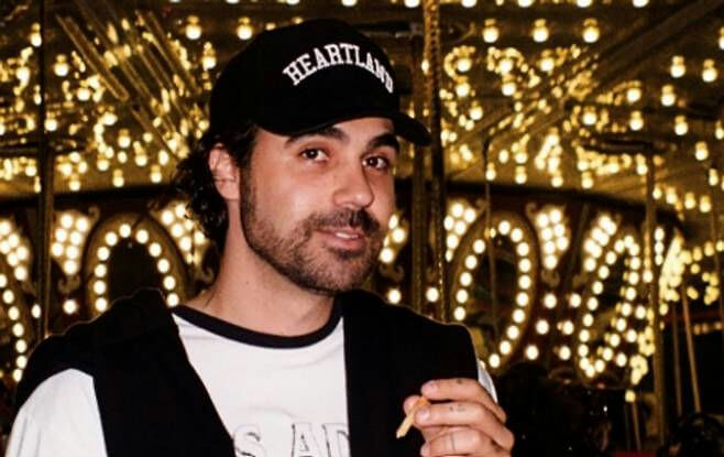 people-1-man-A-black baseball cap with "HEARTLAND" written in white-B-standing in front of a brightly lit carousel or similar attraction-C-holding a small, light-colored object between his fingers