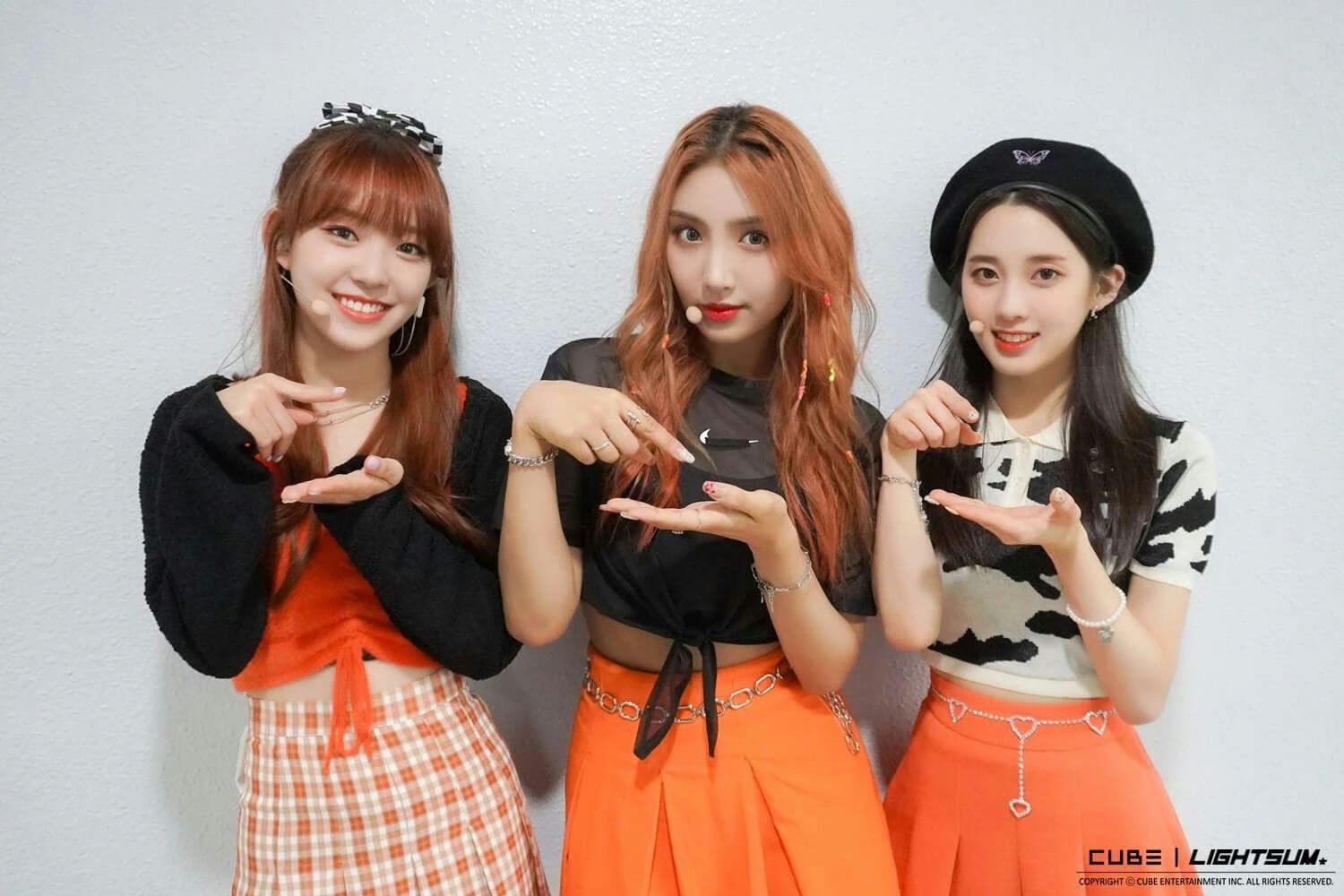 people-three girls-A-K-pop-A-orange clothing-A-black accessories-B-smiling-B-standing close together-C-pointing hands-C-casual setting