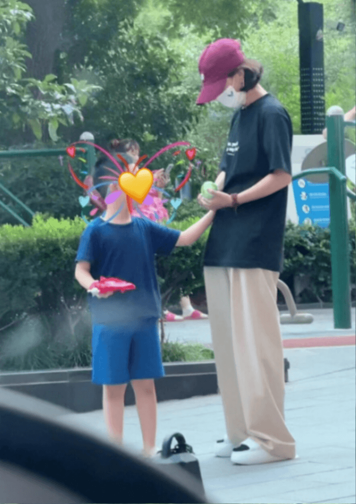 people-woman-burgundy cap-child-blue shirt and shorts-outdoor play area-park-beige wide-leg pants-face obscured by emoji