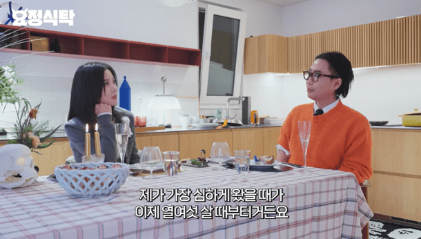 people-woman with dark hair and grey uniform-sitting at a table-man with glasses and orange sweater-having a conversation-indoor setting with kitchen background
