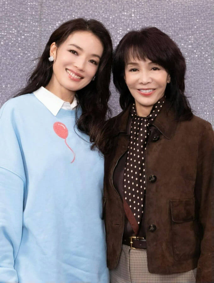 people-Two women-smiling-wearing casual attire-standing side-by-side-light blue sweatshirt with balloon graphic-brown suede jacket with polka dot scarf-against a sparkly silver background