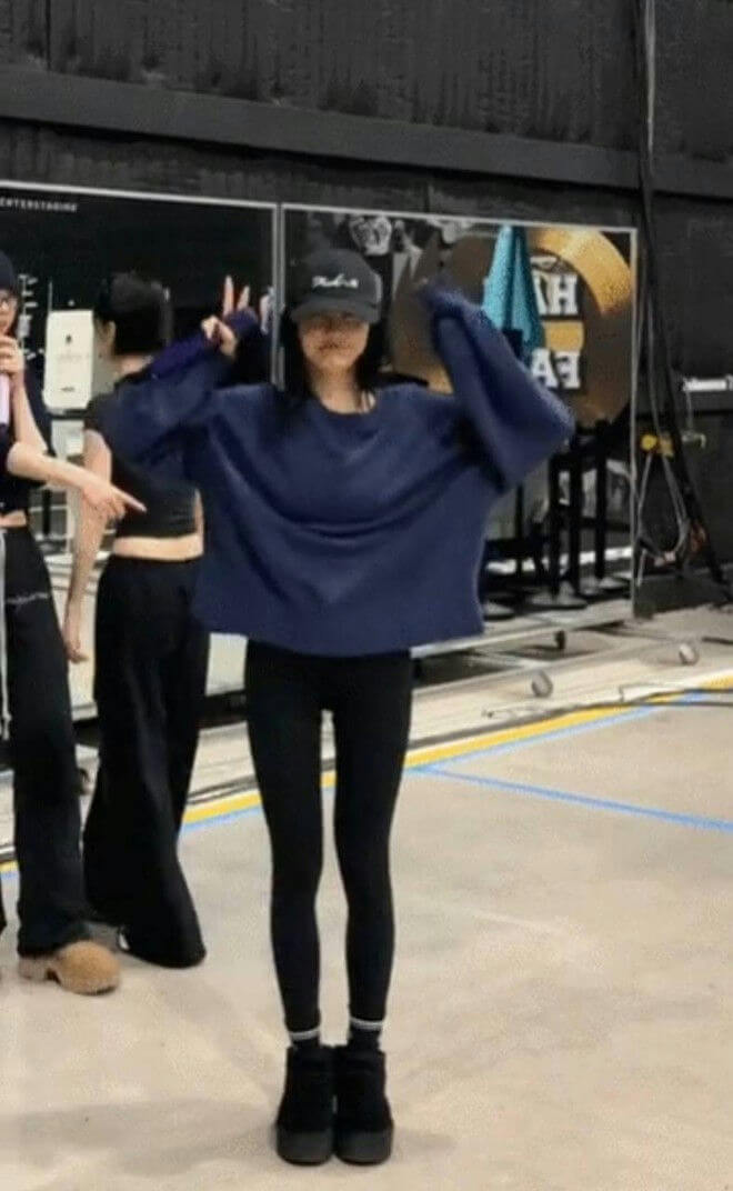people-One person in the center-wearing an oversized navy blue sweatshirt and black leggings-and a black baseball cap-making a peace sign with both hands-Two other people partially visible on the left-indoors or in a studio setting-with visible text/logos in the background
